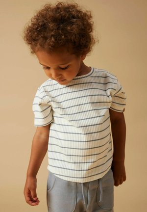 Lille barn med krøllet hår iført en hvid, kortærmet ribstrikket skjorte med vandrette mørke striber, kigger nedad mod en beige baggrund.