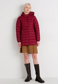 Burgundska puffer jakna s kapuljačom, s horizontalnim šavovima, punim zatvaračem, u kombinaciji s smešnim kordon suknjom i crnim čizmama do kolena.