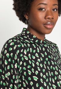 Blusa preta com manchas irregulares verdes e creme, gola alta e textura de tecido suave, com um detalhe de botão na gola.