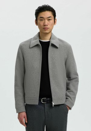 Homme debout, portant une veste en laine gris clair zippée sur un pull noir et un pantalon gris foncé avec une ceinture noire tressée.