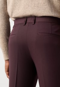 Gros plan d'une personne portant un pantalon violet foncé avec des poches arrière et un pull beige à manches longues rentré.