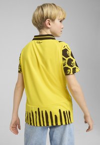 Camiseta deportiva amarilla de manga corta, con un patrón de salpicaduras negras en los hombros y un acento de rayas negras en el dobladillo.