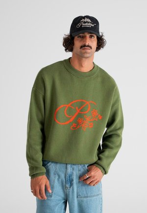 Mann mit Schnurrbart, der eine schwarze Mütze, einen grünen Pullover mit rotem Blumenmotiv "P" und hellblaue Jeans trägt, posiert vor einem weißen Hintergrund.