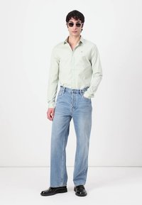 Cămașă verde deschis cu mâneci lungi, blugi albastri cu croială largă și pantofi negri. Model purtând ochelari de soare rotunzi, stând în fața unui fundal alb.