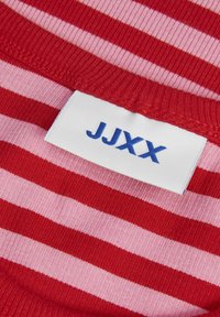 Chemise rayée rouge et rose avec un col côtelé. Comprend une étiquette blanche avec du texte bleu "JJXX" près du col. Texture de tricot lisse.