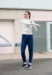 Kobieta w odzieży sportowej rozciąga ramię wzdłuż klatki piersiowej na miejskim chodniku obok metalowej bramy i ściany budynku.