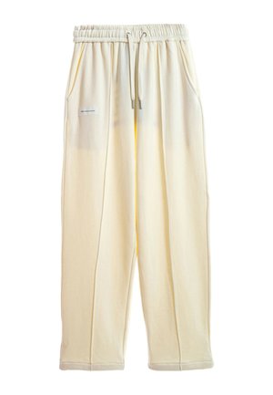 Pantalones deportivos holgados de color crema con cinturilla elástica, cordón, bolsillos laterales y una pequeña etiqueta rectangular en el bolsillo izquierdo.