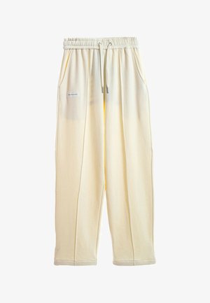 Pantalones deportivos holgados de color crema con cinturilla elástica, cordón, bolsillos laterales y una pequeña etiqueta rectangular en el bolsillo izquierdo.