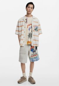 Korte mouwen gestreepte shirt in beige en oranje, graphic t-shirt met palmbomen, beige cargoshorts met kleurrijke prints, en sneakers.