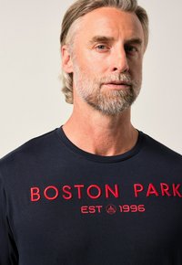 Námořnické bavlněné tričko s kulatým výstřihem, které má červený vyšitý nápis "BOSTON PARK EST 1996". Tričko má hladkou texturu a volný střih.
