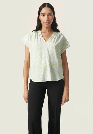 Lyse pastelfarvet bluse med V-hals og korte, vide ærmer, der har et svagt gult og hvidt abstrakt mønster; sat sammen med sorte bukser.
