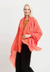 Korallfarbener Schal mit Fransenrändern, leichtes Gewebe, elegant drapiert. Kombiniert mit einem braunen Oberteil und schwarzen Hosen.