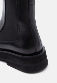 Bottine en cuir noir avec semelle épaisse et panneau élastique latéral, vue de dos et de côté sur fond blanc.