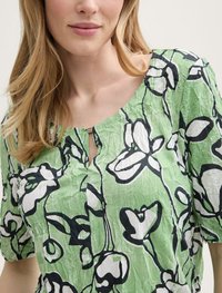 Grüne Bluse mit kurzen Ärmeln, die eine gekräuselte Textur, schwarze Blumenmuster und ein Schlüsselloch-Ausschnittdetail aufweist.