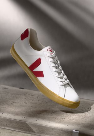 Hvit lær sneaker med røde detaljer og logo. Har rund tå, teksturert gummisåle og flate snøringer. Minimalistisk design.