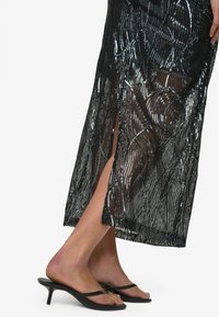 Jupe maxi en sequins noirs avec un motif texturé, fente sur le côté et finition transparente, associée à des sandales à talons noires.