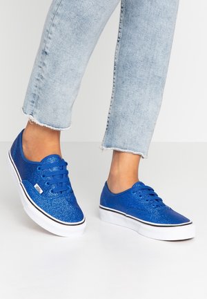 Füße, die glitzernde blaue Vans-Sneaker tragen, kombiniert mit kurz geschnittenen, hell gewaschenen Jeans mit ausgefranstem Saum auf weißem Boden.