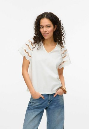T-shirt blanc à manches courtes avec un décolleté en V et un décor en dentelle à bords festonnés sur les manches. Le tissu semble doux et léger. Accompagné de jeans bleus.
