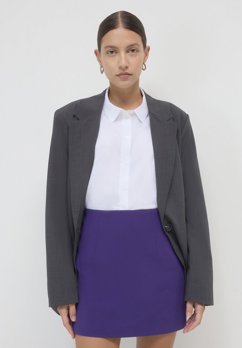 Blazer grigio sopra una camicia bianca con colletto, abbinato a una gonna corta viola. Il blazer ha una texture liscia e un singolo bottone.
