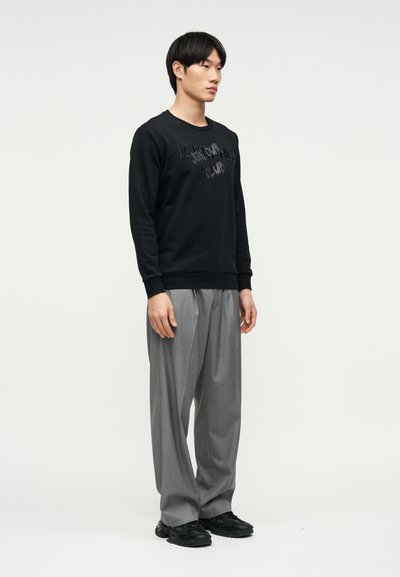 Νέος άνδρας που στέκεται, φοράει μαύρο φούτερ με κείμενο και φαρδιά γκρι παντελόνια, μπροστά σε απλό φωτεινό φόντο.