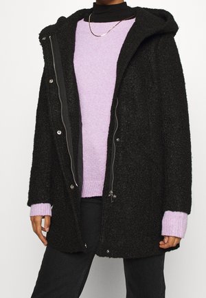 Person iført en sort tekstureret hættejakke over en lavendelfarvet sweater, sort rullekrave, sorte bukser og en tynd guldkæde.