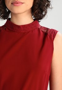 Blouse rouge présentant un col en dentelle haut et des détails en dentelle sur les épaules, en tissu lisse et léger.