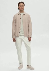 Homem em pé a usar uma camisa-bege clara sobre um sweatshirt branco, calças brancas e sapatos slip-on beges, contra um fundo branco simples.