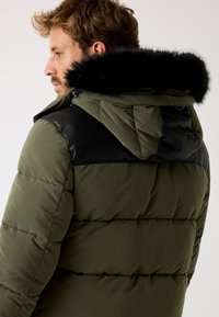 Padded jacket w kolorze oliwkowym z czarnymi skórzanymi akcentami na ramionach i odczepianym czarnym kapturkiem z sztucznego futra. Widoczna pikowana tekstura i zamek błyskawiczny.