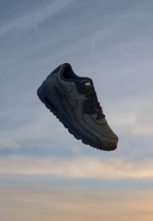 Μονό μαύρο αθλητικό παπούτσι Nike με υφή, να αιωρείται πάνω από ένα συννεφιασμένο ουρανό κατά το ηλιοβασίλεμα.