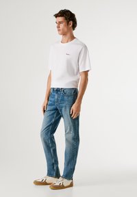 Valge lühikeste varrukatega t-särk, sinised teksad ja beežid tenniseketid. T-särgil on väike broderi logoga. Denimil on kerge kulumine.