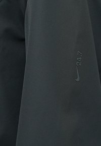 Σκούρο γκρι αθλητικό τοπ με λεία υφή. Διαθέτει διακριτικό λογότυπο Nike και το "24.7" τυπωμένο στο μανίκι. Απλός, λειτουργικός σχεδιασμός.