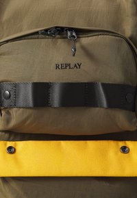 Oliveová zelená batoh s čiernymi komponentmi, horným zipsom a výrazným žltým spodným pruhom; obsahuje logo "REPLAY".