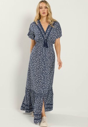 Vrouw die een marineblauwe maxi-jurk met bloemenprint draagt, korte mouwen, V-hals met kwastjes, voorsplit en witte sneakers.