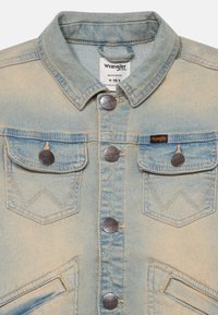 Gaiši zils džinsu žakets ar klasisku apkakli, metāla pogām un divām krūšu kabatām, kas rotātas ar zigzaga šuvju rakstu. Wrangler logo.