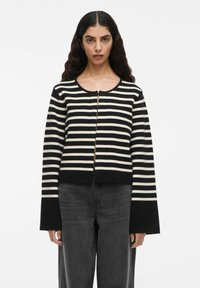 Donna con lunghi capelli scuri e mossi, che indossa un maglione a righe orizzontali bianco e nero con maniche a campana e jeans a vita alta grigio scuro.