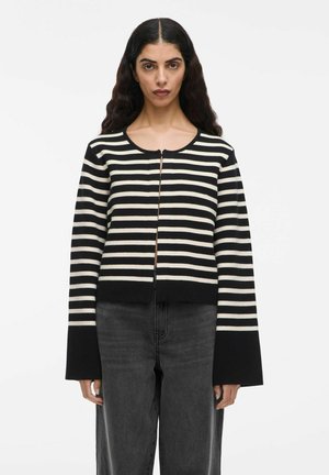 Kvinde med langt mørkt bølget hår iført en sort og hvid vandret stribet sweater med udvidede ærmer og mørkegrå højtaljede jeans.