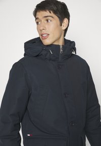 Navy blue hooded jacket with a zip closure, large front buttons, and two side pockets. Features a logo accent in red, white, and blue.