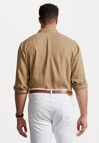 Ljusbrun linneskjorta med uppvikta ärmar, ryggdetalj, i kombination med vita byxor och ett brunt bälte. Slät textur och avslappnad stil.