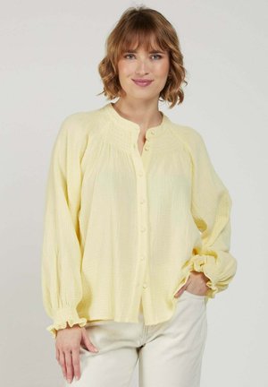 Femme aux cheveux courts et ondulés portant un chemisier ample boutonné jaune pâle et un pantalon clair, debout avec une main dans la poche.