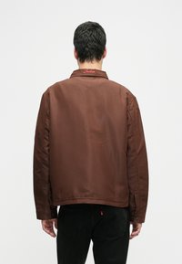 Homem a usar uma jaqueta castanha com "Jacker" bordado em vermelho no colarinho, combinada com jeans pretos, de costas contra um fundo liso.