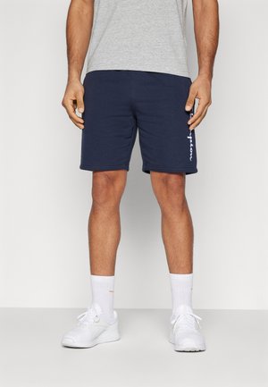 Pantalones cortos deportivos de color azul marino, fabricados con un tejido suave, que cuentan con una cinturilla elástica y texto blanco en la pierna izquierda. Llevados con zapatillas y calcetines blancos.
