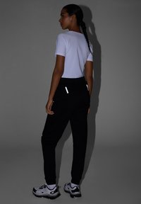 Femme aux cheveux tressés portant un t-shirt blanc, un jogging noir avec des détails réfléchissants et des baskets blanches, debout devant un fond gris.