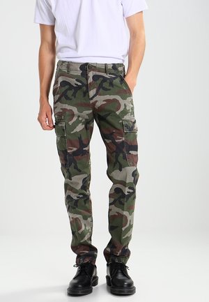 Mężczyzna ubrany w zielone, brązowe i czarne spodnie typu cargo w kamuflażu z bocznymi kieszeniami, białą koszulkę i czarne sznurowane buty, stojący z jedną ręką w kieszeni.