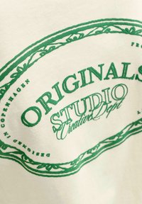 Witte katoenen T-shirt met groene grafische print en de tekst "ORIGINALS STUDIO Creative Dept" met een decoratieve rand. Zachte textuur.