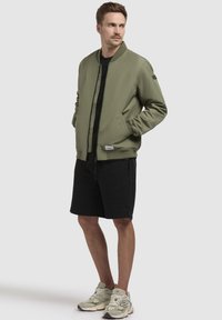 Olivengrüne Bomberjacke mit geripptem Kragen, Reißverschluss vorne und seitlichen Taschen, kombiniert mit schwarzen Shorts und hellfarbenen Sportschuhen.