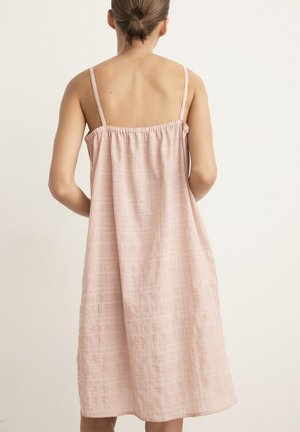 Mujer con cabello castaño recogido en un moño bajo, vistiendo un vestido sin mangas, holgado y de color rosa claro con tirantes delgados, vista desde atrás.