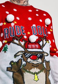 Röd och vit jultröja med en häftig ren med solglasögon, röd mössa, guldhalsband och vita pom-poms på framsidan.