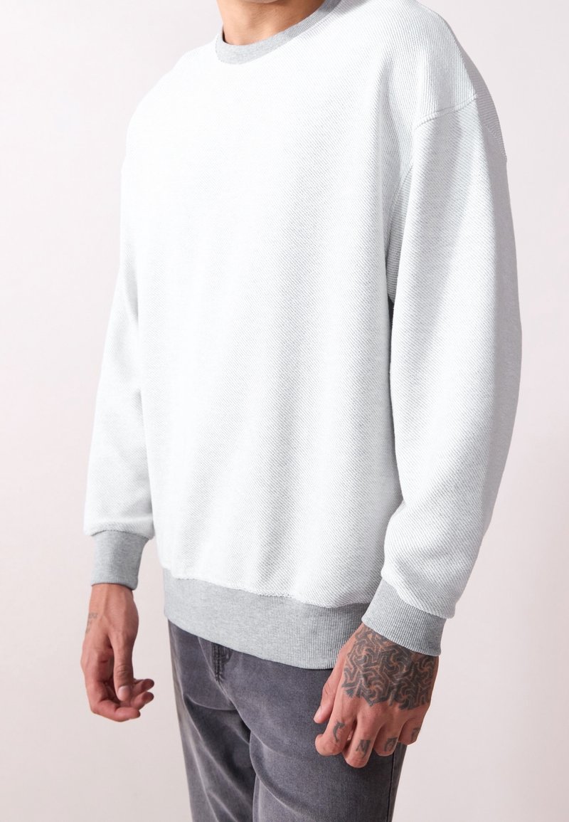 Sweat-shirt gris clair en tissu texturé, col rond et poignets côtelés. Présente un ourlet arrière légèrement plus long et des accents gris.