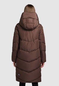 Ruskea toppatakki hupulla, jossa tikatut siksak-kuviot ja pitkät hihat, ulottuu puoliväliin pohjetta. Musta korostelogo helmassa.