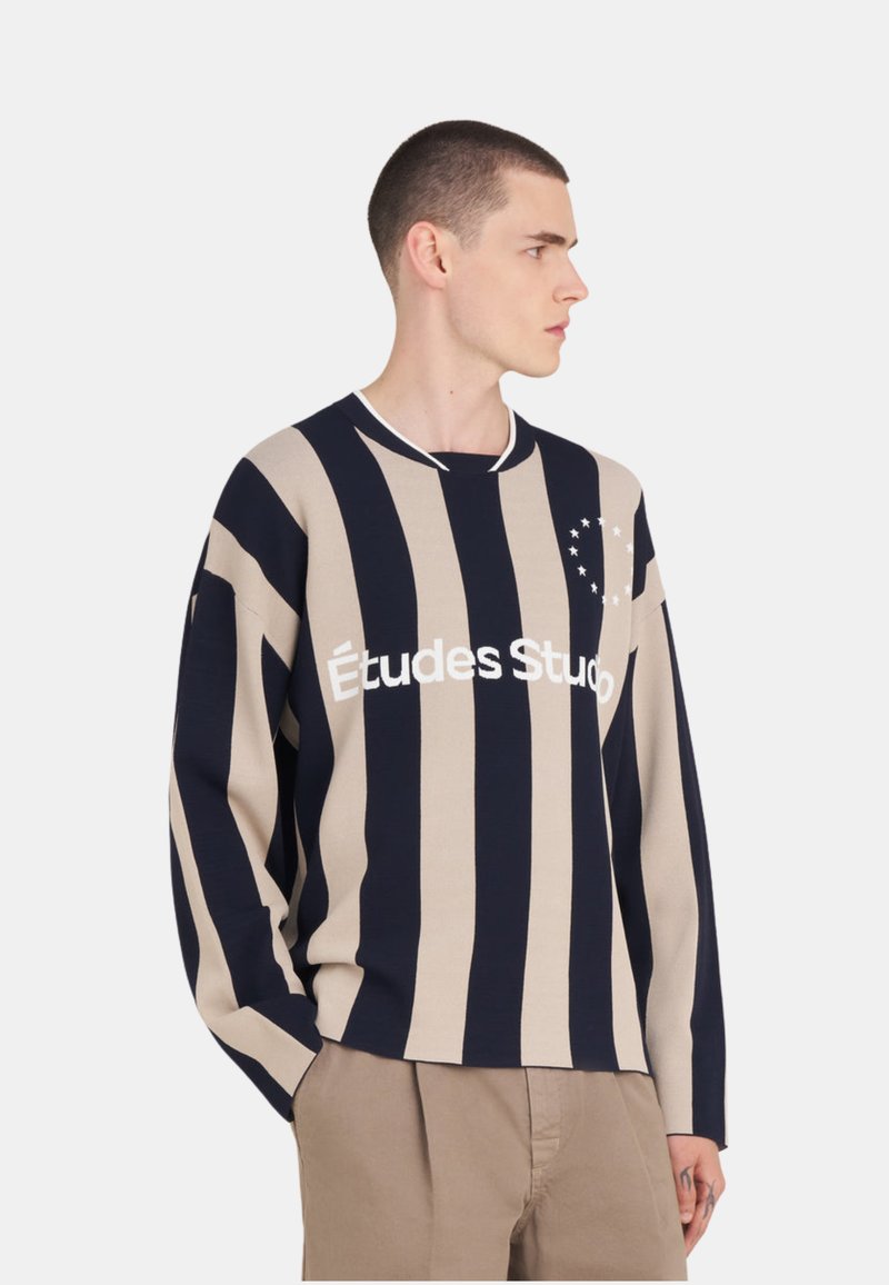 Jeune homme aux cheveux courts portant un pull à rayures verticales beige et noires avec le texte "Études Studio", associé à un pantalon beige.